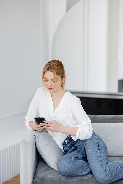 Blonde woman in white blouse and jeans using smartphone and listening music in earphone on couch