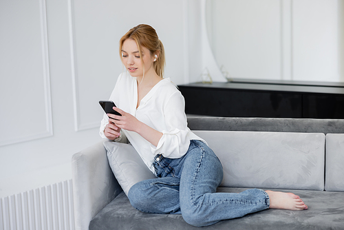 Barefoot woman in earphone using mobile phone on couch at home