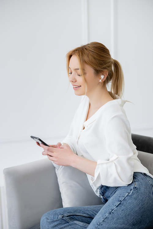 Positive young woman in earphone using smartphone in living room