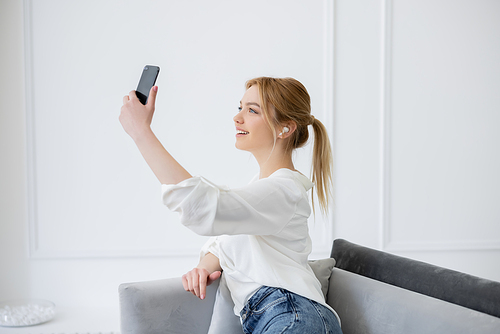 Positive young woman in earphone having video chat on mobile phone on couch