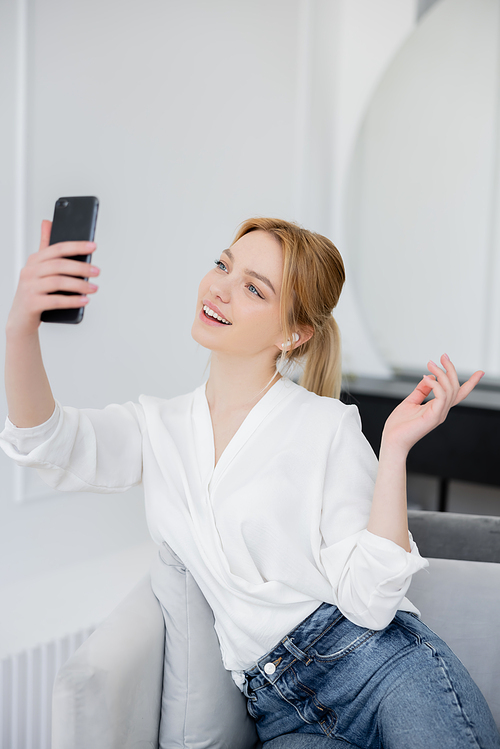 Positive young woman in blouse and earphone having video call on smartphone in living room