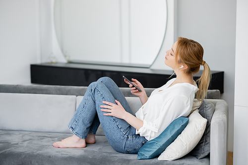 Side view of young woman in earphone holding cellphone on couch at home