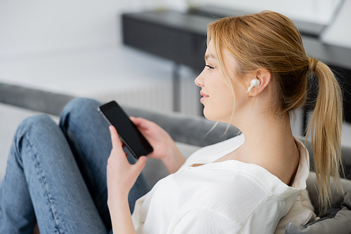 Side view of pretty blonde woman in earphone holding blurred smartphone at home