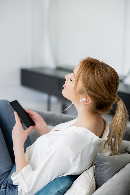Side view of blonde woman in blouse and earphone holding mobile phone on couch