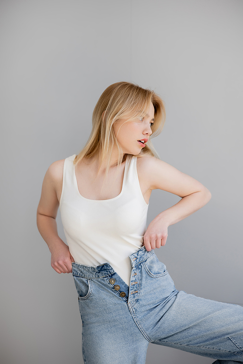 Blonde woman in bodysuit and jeans looking away isolated on grey