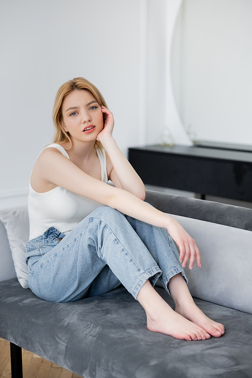 Barefoot woman in jeans looking at camera while sitting on couch at home