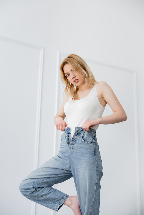 Young woman in bodysuit touching jeans and looking at camera in living room
