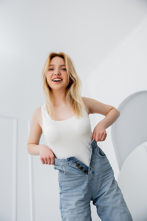 Low angle view of positive blonde woman touching jeans at home