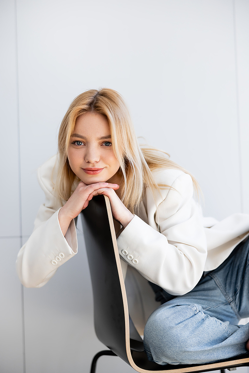 Portrait of blonde woman in jacket looking at camera on chair at home