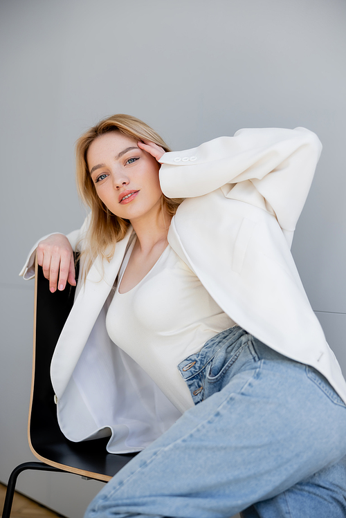 Trendy woman in jeans and jacket looking at camera on chair