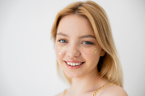 Cheerful blonde woman with glitter makeup looking at camera isolated on grey