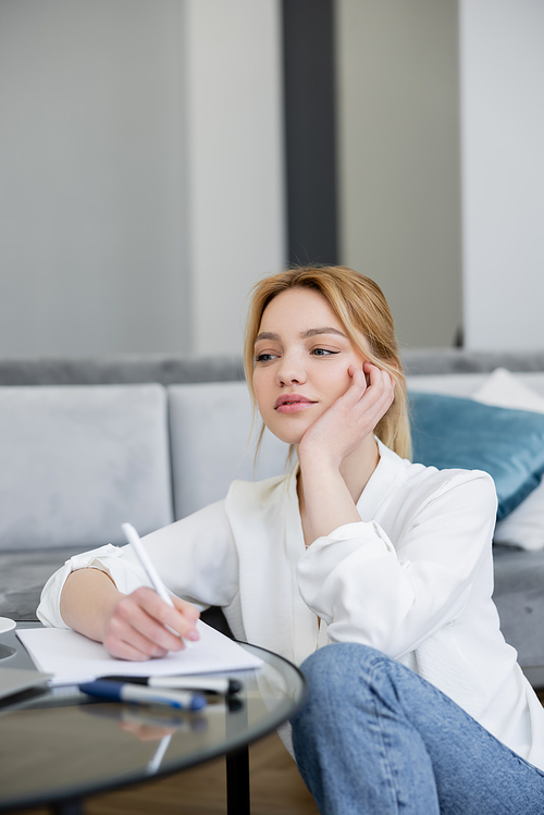 Pensive woman in blouse writing on notebook on coffee table