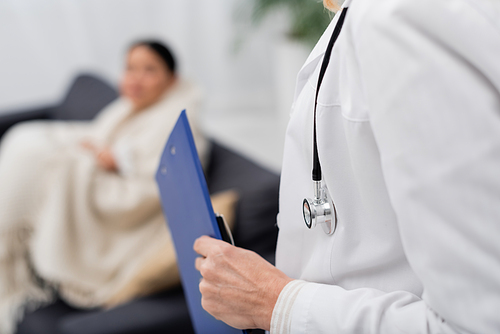 Cropped view of doctor in white coat holding clipboard near blurred patient at home