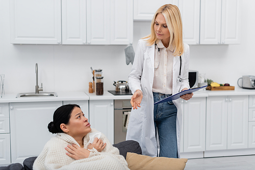 Mature doctor holding clipboard while talking to sick asian patient at home