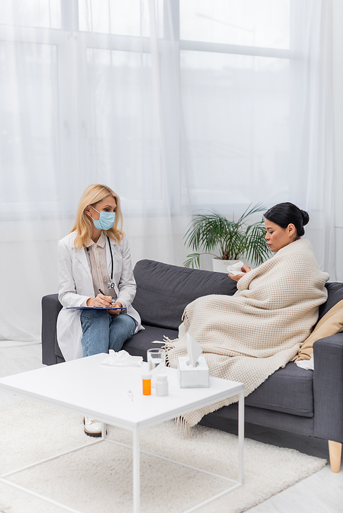 Doctor in medical mask writing on clipboard near sick asian patient at home