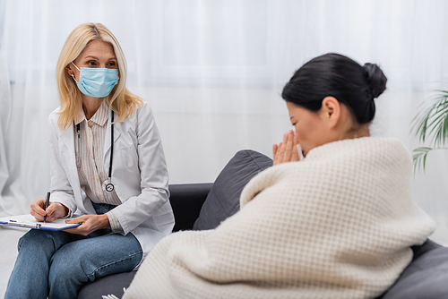 Doctor in medical mask holding clipboard near blurred asian patient at home