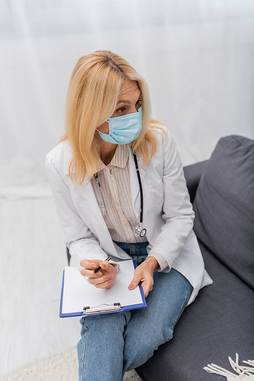 Mature doctor in medical mask holding clipboard and sitting on couch