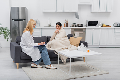 Asian woman in blanket holding napkin near doctor in medical mask writing on clipboard at home