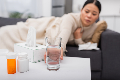 Blurred woman taking glass of water near pills and napkins at home