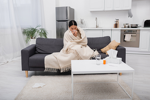 Diseased asian woman holding napkin near pills on table at home