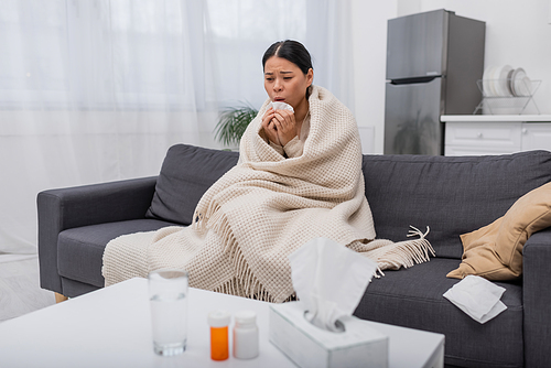 Tired and sick asian woman holding napkin near blurred pills at home