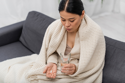 sick asian woman in blanket holding glass of water and pills at home