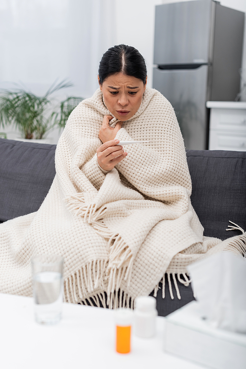 Worried asian woman in blanket looking at thermometer near blurred pills and napkins at home