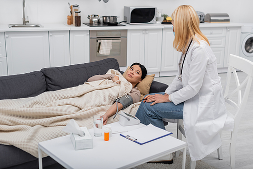 Sick asian patient lying near doctor in medical mask and tonometer at home