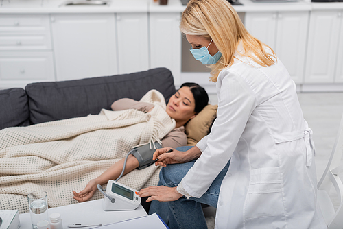 Doctor in medical mask looking at tonometer near blurred asian patient at home