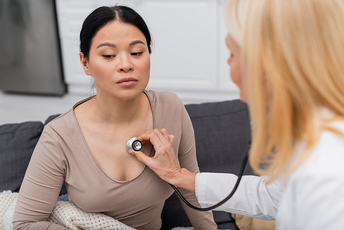 Blurred doctor with stethoscope checking lungs of sick asian woman at home