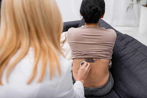 Back view of blurred doctor holding stethoscope near back of patient at home