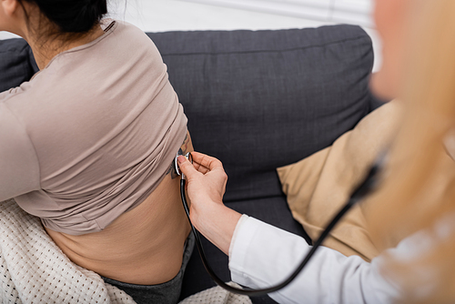 Cropped view of blurred doctor with stethoscope checking back of woman at home