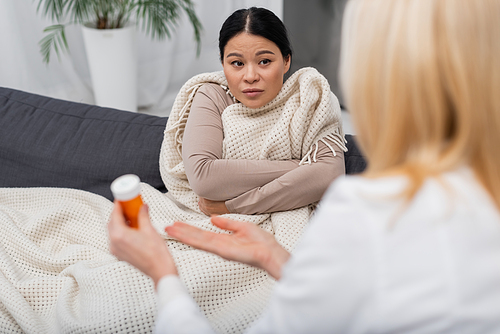 Sick asian woman in blanket looking at blurred doctor pointing at pills at home