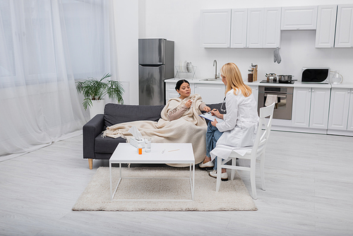 Sick asian woman holding napkin and pointing with finger on clipboard near doctor at home