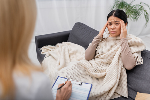 Asian woman holding napkin and suffering from headache near blurred doctor with clipboard at home