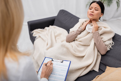 Blurred doctor writing on clipboard near sick asian woman touching neck at home