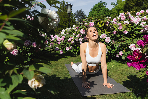 laughing woman with closed eyes practicing cobra pose on yoga mat near blossoming plants