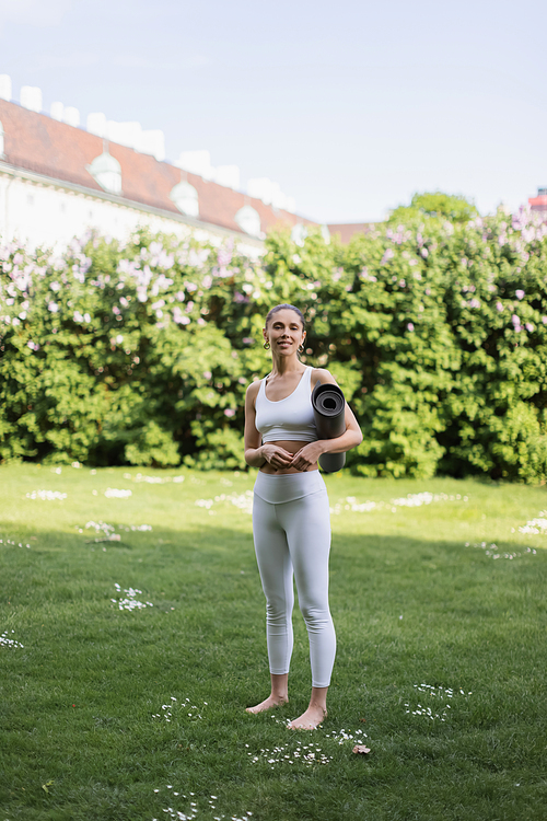 full length of barefoot woman in white sportswear standing with yoga mat on green lawn