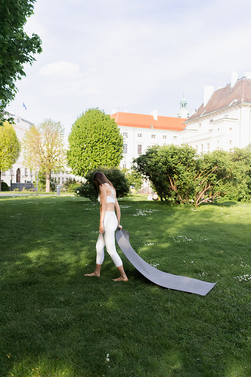 barefoot woman in white sportswear holding yoga mat in city park