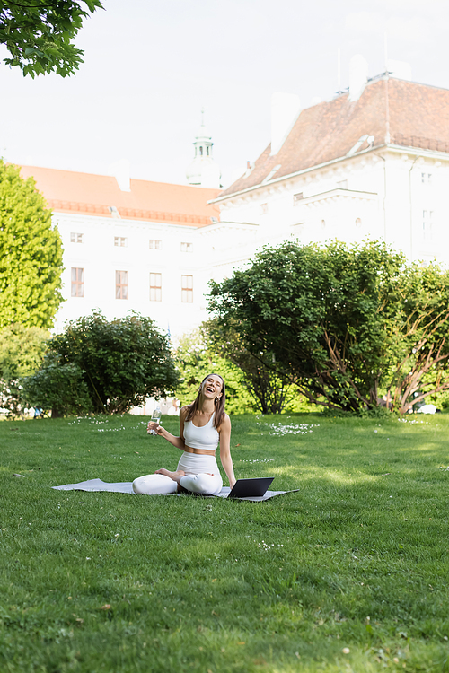 laughing woman with sports bottle sitting in lotus pose near laptop in park