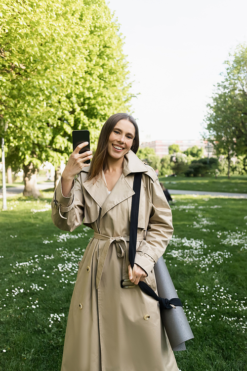 happy woman in trench coat standing with fitness mat and taking selfie on smartphone in park