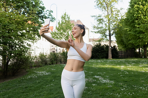 cheerful sportswoman sticking out tongue and showing victory sign while taking selfie in park