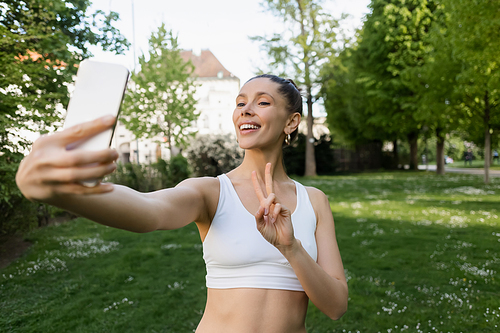 cheerful woman in white sports bra showing victory gesture and taking selfie on blurred smartphone in park