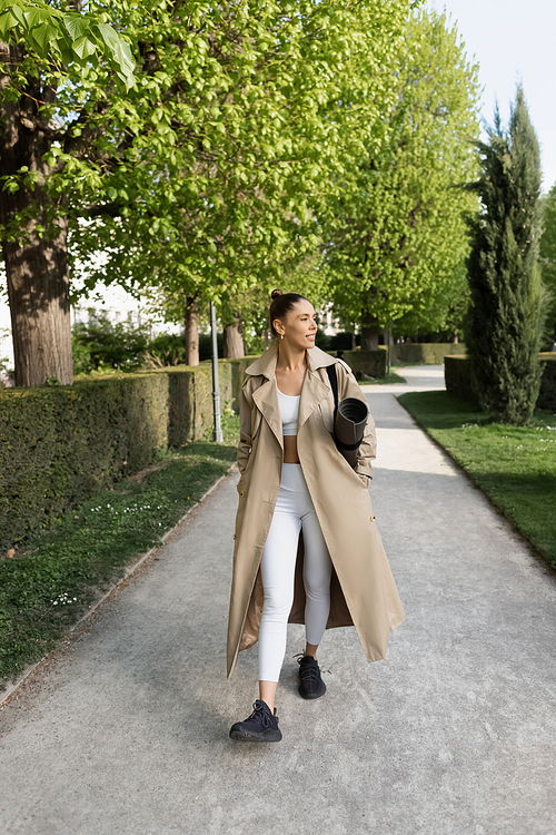 full length of smiling sportswoman in trench coat walking with fitness mat in park