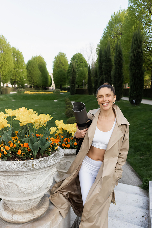 cheerful woman in sportswear and trench coat holding fitness mat near park vases with flowers