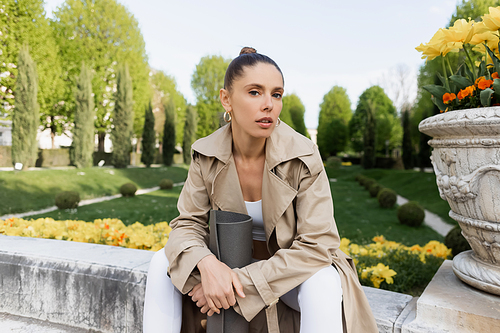 woman in trench coat looking at camera while sitting with fitness mat near park vase