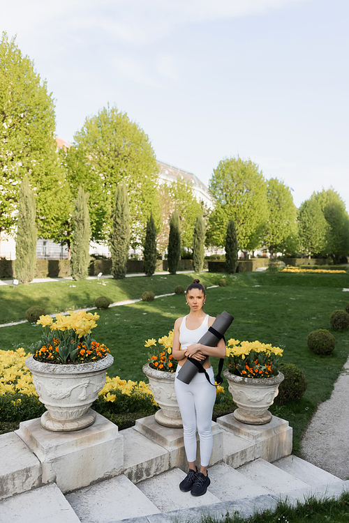 full length of sportswoman holding fitness mat while standing on stairs near park vases