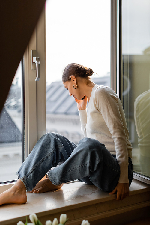 barefoot woman in jeans talking on mobile phone and looking down through window