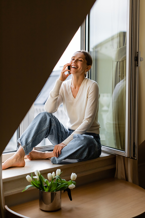 cheerful barefoot woman in jeans sitting on windowsill near tulips and talking on mobile phone