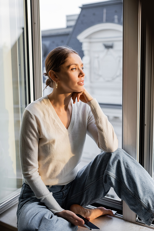 dreamy woman looking away while sitting on windowsill at home